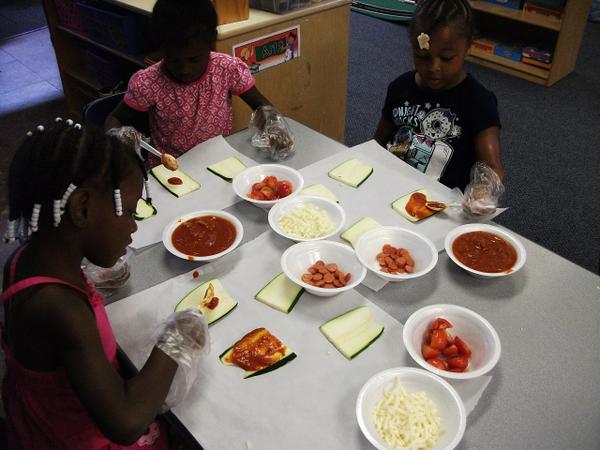 Children sit at a table with slices of eggplant and bowls containing cheese, sliced tomatoes, and tomato sauce.